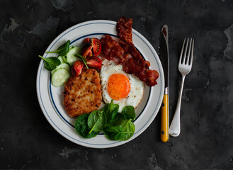 Delicious breakfast - fried egg, bacon, potato salad, vegetables, spinach in a plate on a dark background, top view