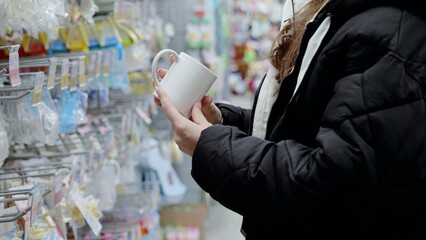 Woman carefully considering a plain white mug while shopping for kitchenware in a supermarket aisle