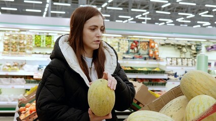 Young lady carefully selecting a ripe and high quality melon in the produce section of a supermarket