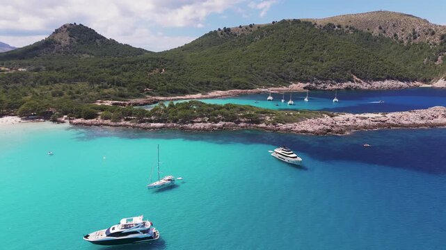 Aerial Drone Panning Shot Over Anchored Boats In Cala Agulla Bay Towards Pine Forest Mountains, Mallorca, Spain.