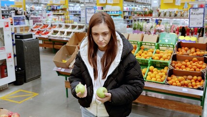 Young woman comparing two green apples while shopping for groceries in the produce section of a supermarket, surrounded by fresh fruits and vegetables
