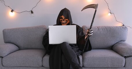 A male actor in a black skeleton suit sits on a sofa with a laptop. Online Halloween celebration, dakrnet, social media. Concept symbol of death with a scythe, grim reaper scythe 