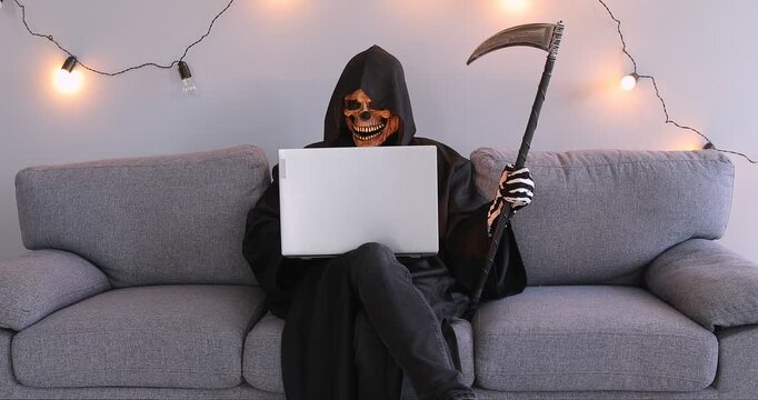 A male actor in a black skeleton suit sits on a sofa with a laptop. Online Halloween celebration, dakrnet, social media. Concept symbol of death with a scythe, grim reaper scythe 