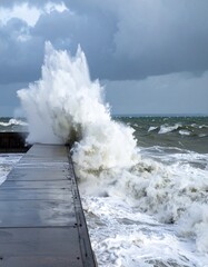 A powerful ocean wave crashes against a dock during bad weather. The scene is dramatic, with rough, churning waters and foamy spray splashing high onto the wooden or concrete dock structure.