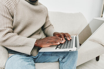 Naklejka premium A young Black man working on a laptop while seated on a cozy couch, wearing a beige sweater and casual jeans, depicting a relaxed and productive home environment
