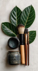 Dark-toned makeup brushes and container atop a light gray surface, accented by lush green leaves