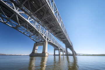 Massive steel truss bridge spans a calm river under a bright blue sky, viewed from below. Industrial architecture and structural detail highlight engineering design. ai generative