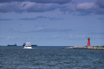 Boats cruising near red lighthouse and ships in calm waters under a cloudy sky