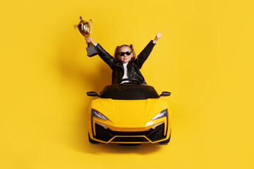 Young girl in black leather jacket and sunglasses sitting in a yellow toy sports car, raising a golden trophy and celebrating against yellow background.