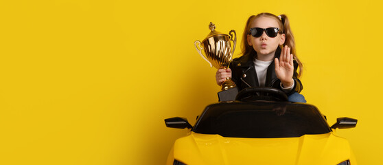 Young girl in black leather jacket and sunglasses sitting in a yellow toy sports car, holding a golden trophy and gesturing stop against yellow background.