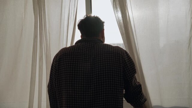Man looking out window, back shot, peeking head between white curtains. Sun shining through curtains. Looking down at city street. Hiding and trying to avoid someone or something. Scared or curious