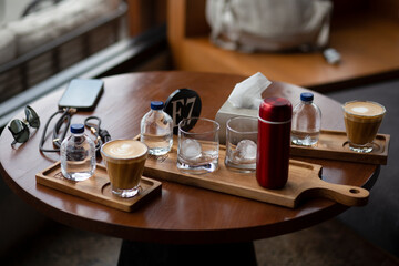 Red compact thermos, two glass tumblers with ice, and a glass of cappuccino arranged on a round wooden table