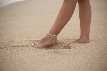Close-up photo of elegant female legs resting on golden sandy beach, adorned with minimalist ankle bracelets