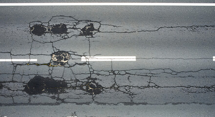 Close up aerial view of a damaged asphalt road surface showing cracks potholes and repair patches indicating wear and tear on infrastructure