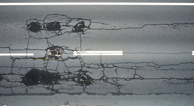 Close up aerial view of a damaged asphalt road surface showing cracks potholes and repair patches indicating wear and tear on infrastructure
