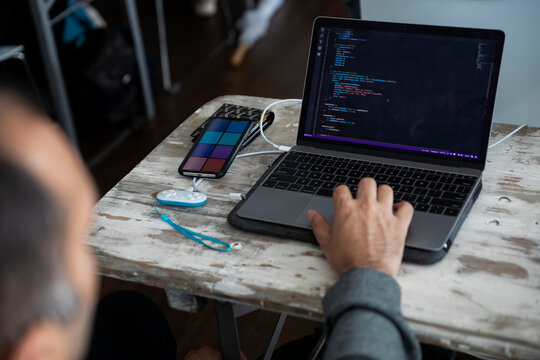 Developer programming on laptop with code editor visible alongside smartphone and charging cables - Powered by Adobe