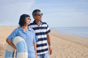 Cheerful senior couple traveler wearing a casual shirt and holding swimming ring in front of the morning Beach with copy space.