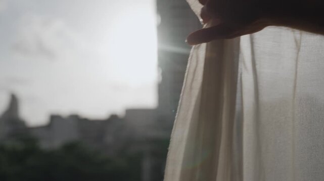 Hand reveals sun behind window curtains in early morning or sunset hour in city. Man pulls fabric on summer day outside. Sun light shines strong with flare. Silhouette of arm and hand indoors