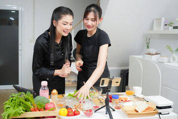 Two asian women cooking food, making breakfast and salad while using smart phone or digital tablet, streaming live video clip or taking selfie photo together with happiness and fun in kitchen at home	