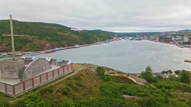 A drone hovers above Brigus South harbour, capturing the wharf with a tall mast, calm waters, fishing boats, and a colorful hillside community along Newfoundland coast.