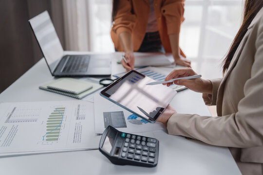 Businesswomen collaborating on financial analysis with a digital tablet, calculator, and laptop while reviewing documents with charts and graphs on the table