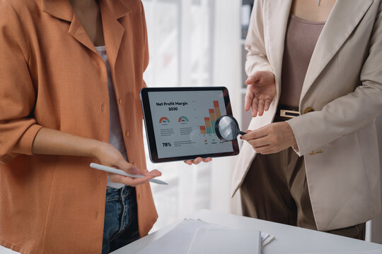 Two businesswomen analyzing financial charts on a digital tablet, using a magnifying glass to discuss growth and profit margins in a collaborative office setting