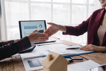 Two businesswomen shaking hands after successful real estate deal in modern office, small wooden house model on foreground, laptop with charts on background