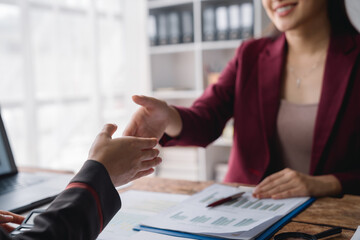 Two businesswomen engaged in a handshake after successfully closing a deal in the office, surrounded by documents and a laptop on the table, symbolizing partnership and collaboration