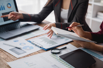 Two businesswomen are discussing financial charts and graphs displayed on a laptop, using a notebook and a pen to analyze data and make informed decisions