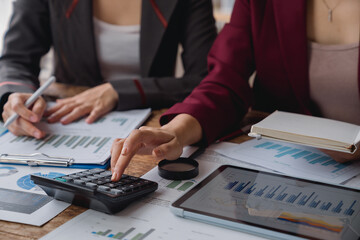 Two businesswomen collaborating in an office, analyzing financial charts and graphs, using a calculator, and taking notes during a focused and productive meeting