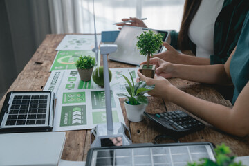 Two environmental engineers are discussing green logistics using a miniature wind turbine and solar panel, analyzing data on a tablet and calculating costs on a wooden table
