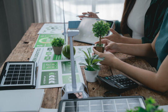Two environmental engineers are discussing green logistics using a miniature wind turbine and solar panel, analyzing data on a tablet and calculating costs on a wooden table