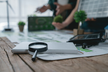 Magnifying glass, solar panel and wind turbine models are lying on a wooden desk while engineers are analyzing a sustainable development project in the background
