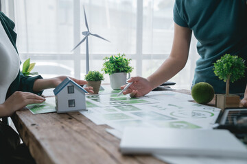 Two architects are discussing an eco-friendly house project, analyzing blueprints and a miniature...