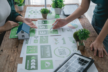 Two businesswomen discussing an environmentally sustainable ESG project, analyzing charts and graphs on carbon emissions and net zero goals, with a miniature house model