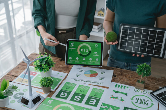 Two businesswomen are using a tablet showing esg icons and holding a photovoltaic panel during a meeting about sustainability and environmental, social and governance - Powered by Adobe