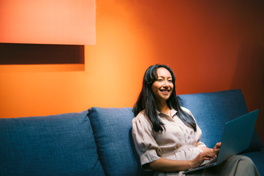 Smiling woman using laptop on sofa indoors with orange wall