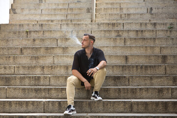 Young, brunette, Hispanic, modern man in black crochet shirt and jeans, smoking sitting in the...