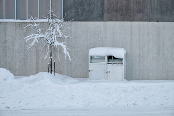 A mailbox stands buried in snow beside a leafless tree against a gray concrete wall during winter.