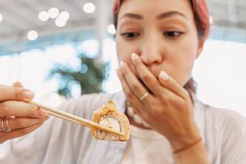 Young woman is holding a sushi roll with chopsticks and covering her mouth, expressing disgust