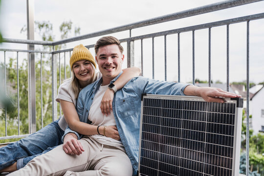 Happy young man and woman sitting with solar panel on balcony