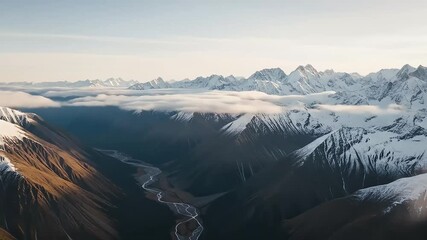 Aerial view of snow-capped mountains with dramatic clouds hugging peaks as a winding river flows through deep valleys under soft golden light at sunrise - Powered by Adobe