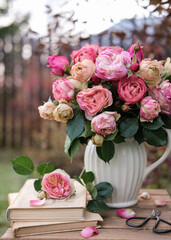Stunning bouquet of pink and peach roses in white pitcher sits on wooden table with old books and garden shears, set against blurred outdoor background. Romantic, rustic aesthetic