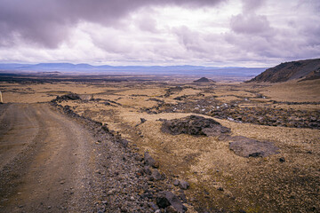 A gravel road winds through Iceland’s barren volcanic desert with dark lava rocks scattered across golden plains beneath heavy clouds
