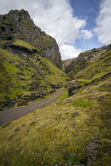 Obraz premium Narrow path winding through moss-covered volcanic cliffs and rugged green valley under a partly cloudy sky in Iceland