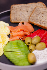 Healthy breakfast plate with scrambled eggs, smoked salmon, sliced avocado, green olives, tomato and whole grain bread. Balanced nutritious meal, close up food photography for menu or diet concept