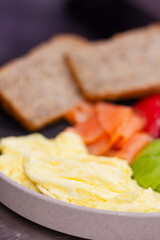 Healthy breakfast plate with scrambled eggs, smoked salmon, sliced avocado, green olives, tomato and whole grain bread. Balanced nutritious meal, close up food photography for menu or diet concept