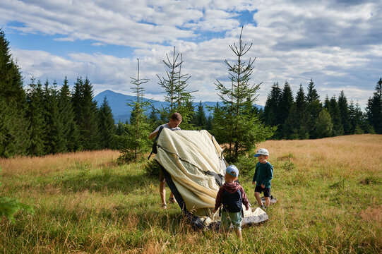 Man hiker and two young boys work together to set up tourist tent on grassy hillside. Pine trees and distant mountains under partly cloudy sky.