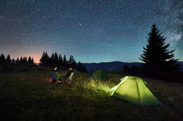 Silhouette of couple hikers resting by campfire under star-studded sky, near illuminated tents at campsite. Night camping in mountains, night sky create magical and peaceful atmosphere.
