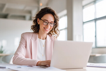Mujer de negocios de 40, con chaqueta rosa, de traje, blusa blanca, gafas graduadas, sentada en una mesa dde despacho trabajando con el ordenador, con gran sonrisa en oficina luminosa con ventanales